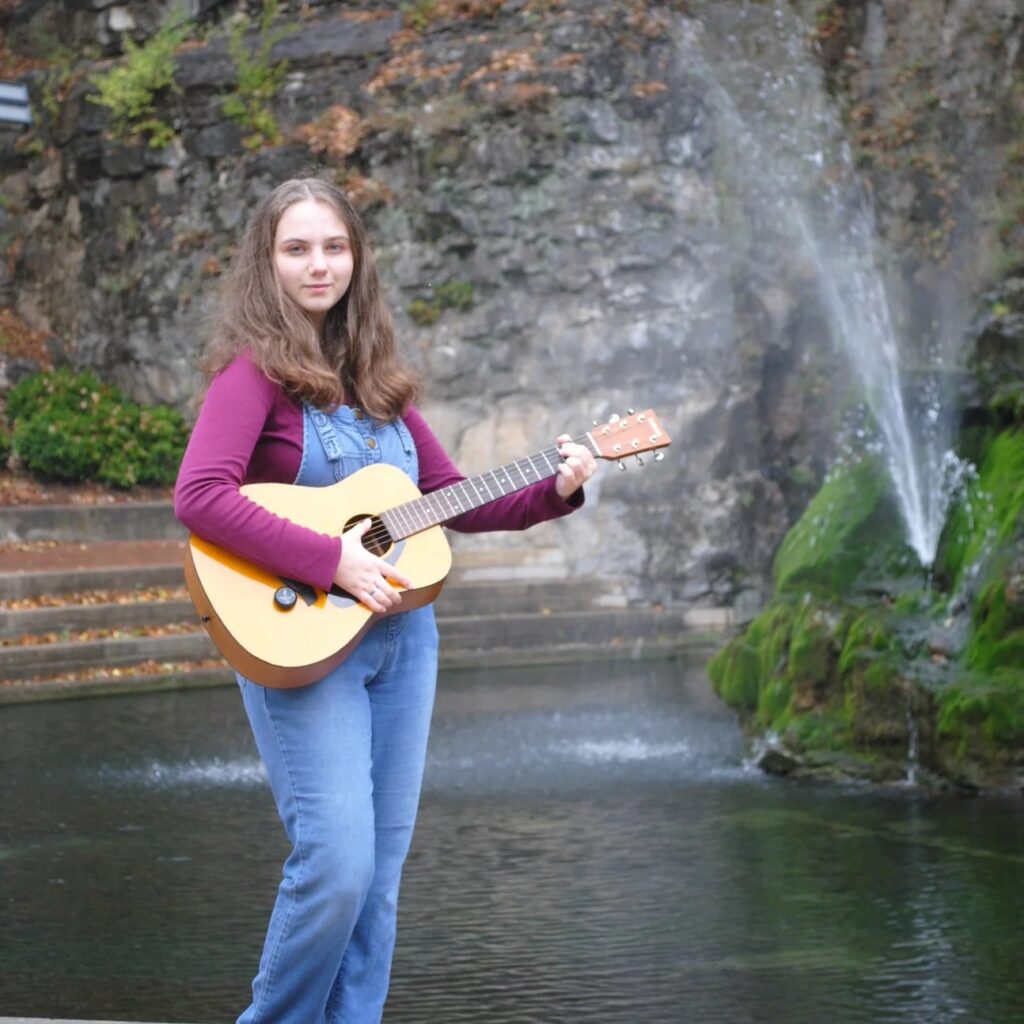 Uncle Emmington is holding a guitar standing in front of a natural spring