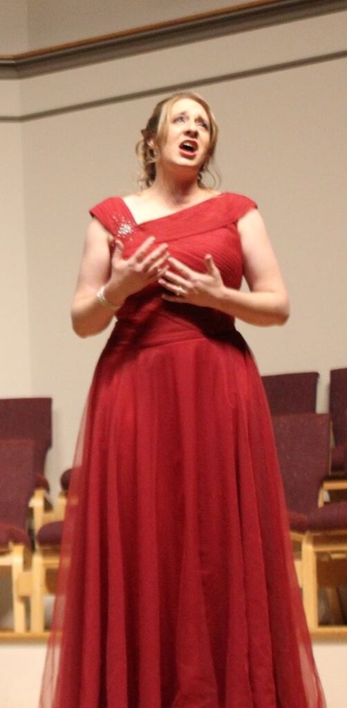 Image of a White woman with a floor-length red dress. The woman is singing.