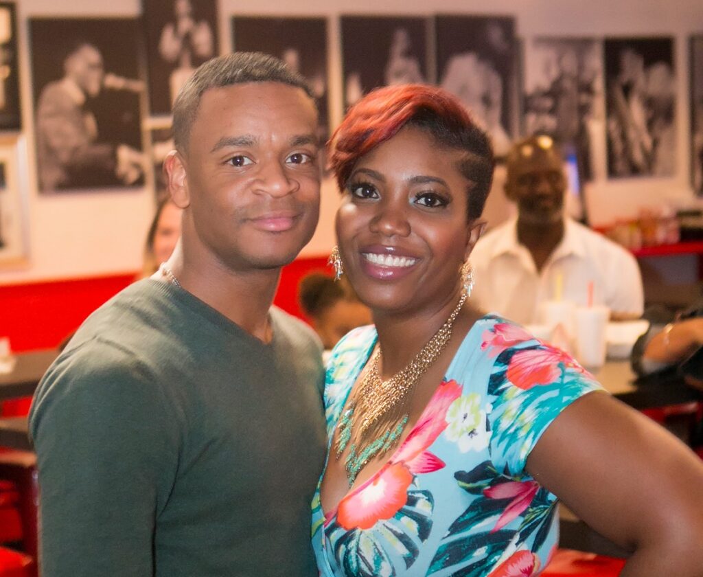 Photo of a Black and a Black woman standing side-by-side and smiling at the camera. They are Charles and Tanya Harris.