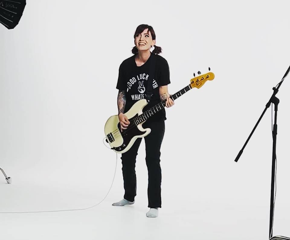 A White woman is holding a bass guitar in a white room, smiling at the camera.