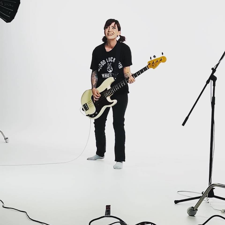 A White woman is holding a bass guitar in a white room, smiling at the camera.