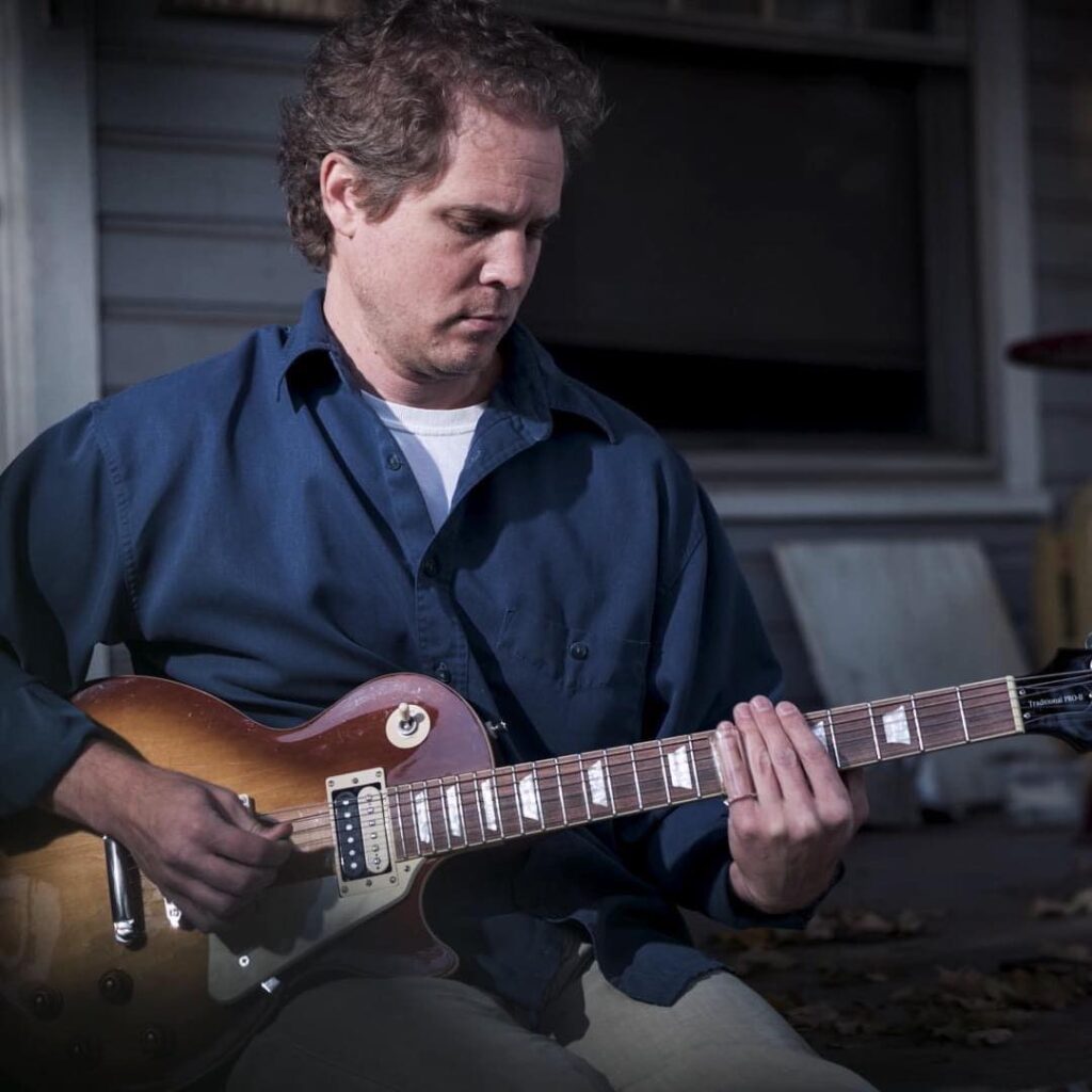 Image of a white man in a blue shirt holding and playing a guitar.